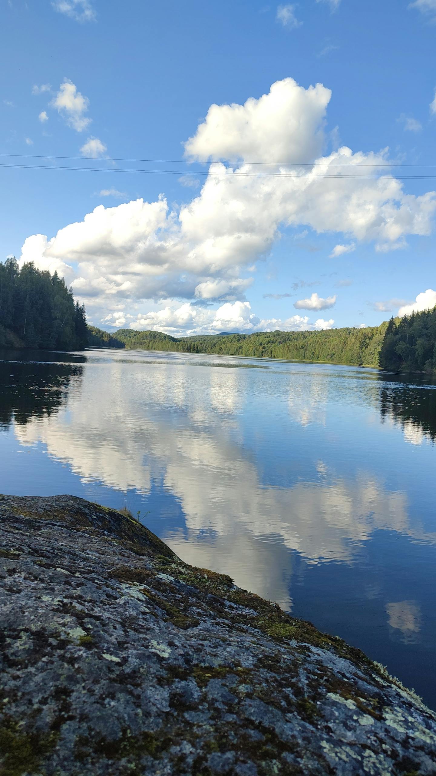 Tranquil lake with fluffy clouds reflected in clear water, surrounded by lush forests.